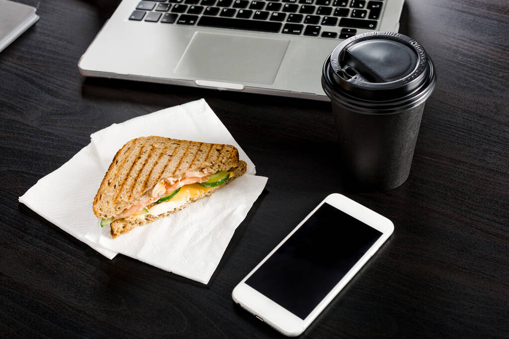 Caffè e un panino sdraiato sulla scrivania accanto al telefono e al portatile. Uno sfondo scuro. Pranzo di lavoro durante il processo di lavoro
. - Foto, immagini