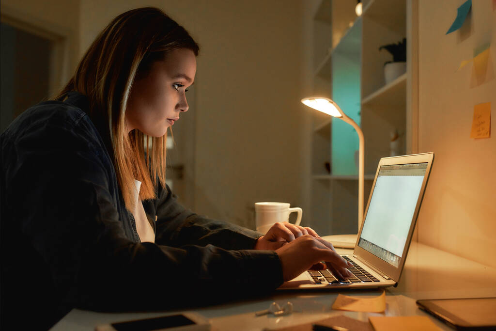 Przepracowuję. Widok z boku młodej białej kobiety pracującej w nocy w domu, używającej laptopa i pijącej kawę. Siedzi w swoim biurze, pracuje zdalnie. - Zdjęcie, obraz