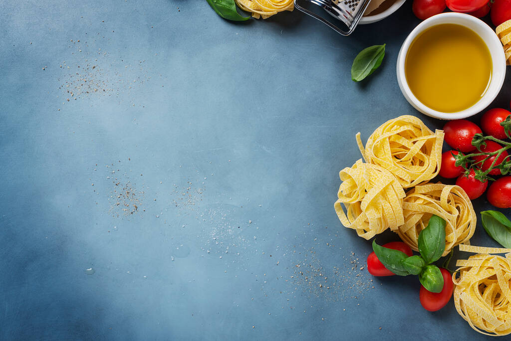 Konzept der italienischen Küche mit rohen Tagliatelle, Tomate, Basilikum, Olivenöl, Salz und Pfeffer auf blauem Grund. Bild von oben mit Kopierraum - Foto, Bild
