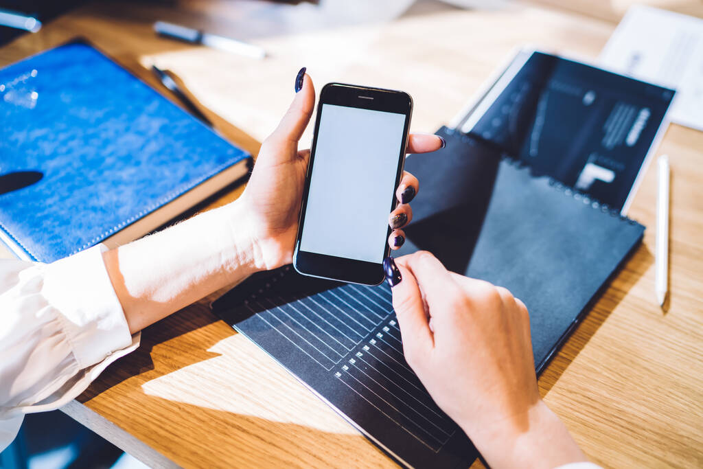 Van boven vrouw met blauwe manicure surfen mobiele telefoon zitten aan houten tafel met documenten notebook in kantoorruimte  - Foto, afbeelding