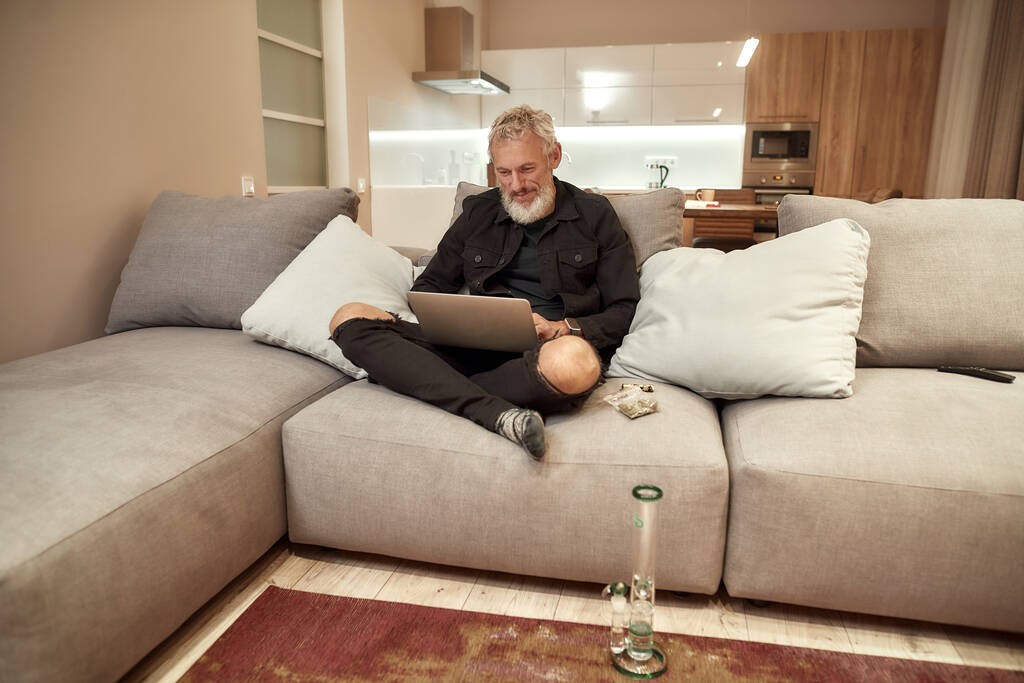 A trabalhar dentro de casa. Homem de meia-idade barbudo usando laptop enquanto sentado no sofá. Bong ou tubo de água de vidro, botões em saco plástico e isqueiro perto dele. Cannabis e conceito de legalização de ervas daninhas
 - Foto, Imagem