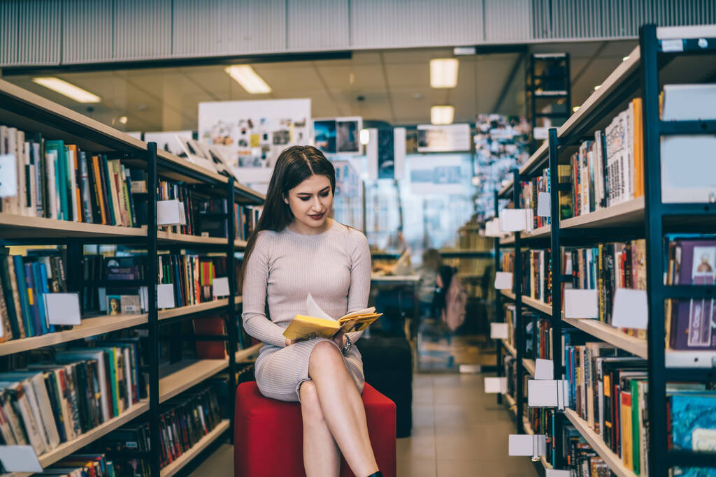 Schöne junge Frau in beigem Kleid sitzt auf Hocker und liest Buch mit gelbem Einband, während sie neben Bücherregalen in der Universitätsbibliothek sitzt - Foto, Bild