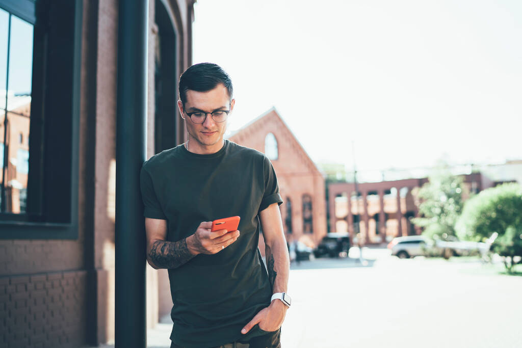 Soustředěný student mužského pohlaví ve stylovém neformálním oblečení a brýlích, opírající se o hromadu při smskování s přáteli na mobilním telefonu v City Street - Fotografie, Obrázek