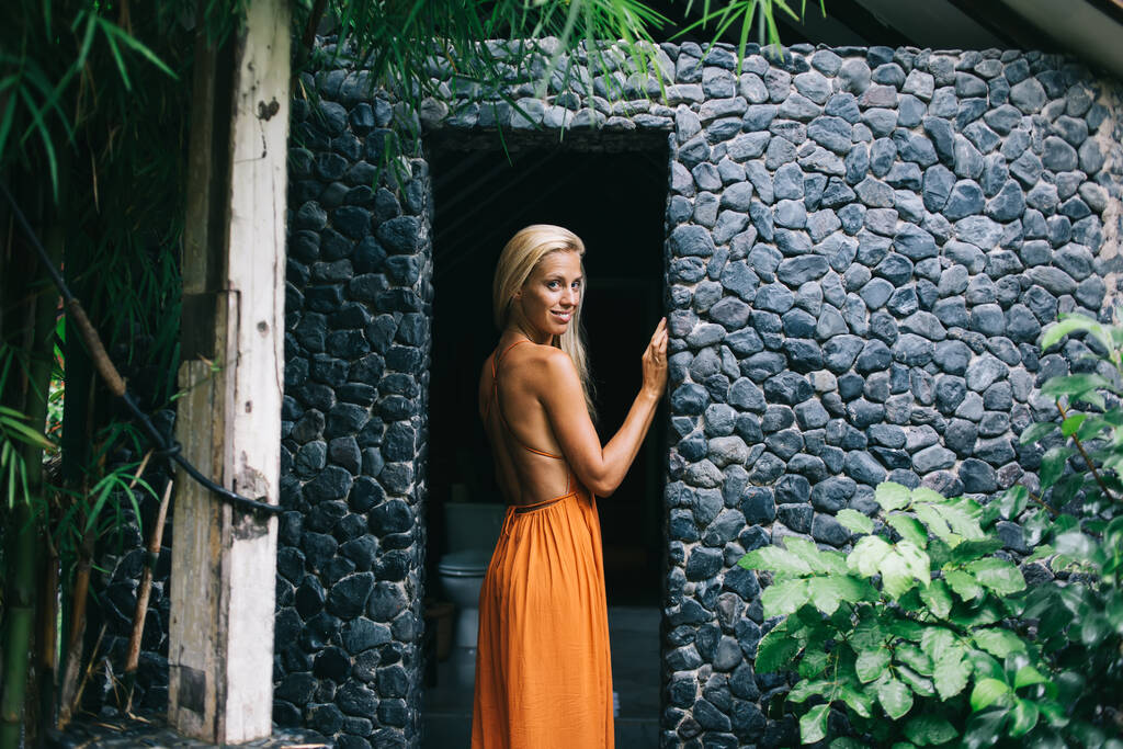 Vista lateral de la hermosa mujer joven en vestido elegante de pie junto a la pared de piedra de piedra de edificio rural exótico con inodoro en Bali - Foto, Imagen