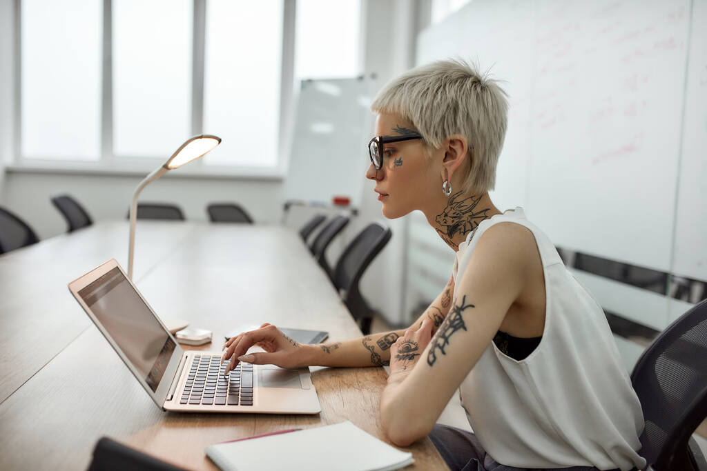 Az üzletasszony a munkára koncentrál. Oldalnézet fiatal és gyönyörű tetovált nő rövid frizurával laptoppal, miközben ül a munkahelyén a tárgyalóban - Fotó, kép