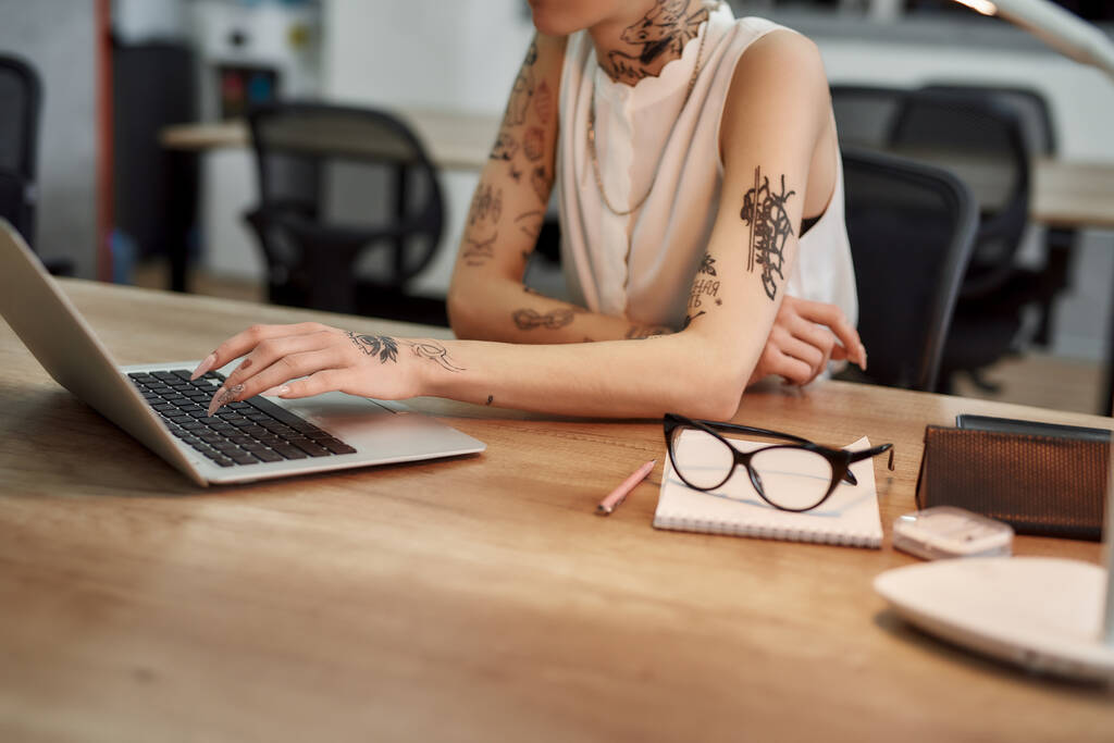 Revisando correo electrónico. Foto recortada de una joven mujer tatuada escribiendo algo en el portátil mientras está sentada en su lugar de trabajo en la oficina moderna
 - Foto, imagen