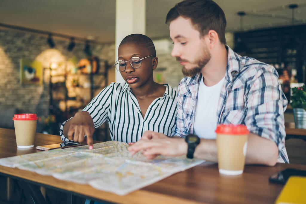 カジュアルな多人種カップルは、旅行先を選択するカフェで紙コップからテーブルドリンクコーヒーに座って、地図で指を指して行く場所を議論 - 写真・画像