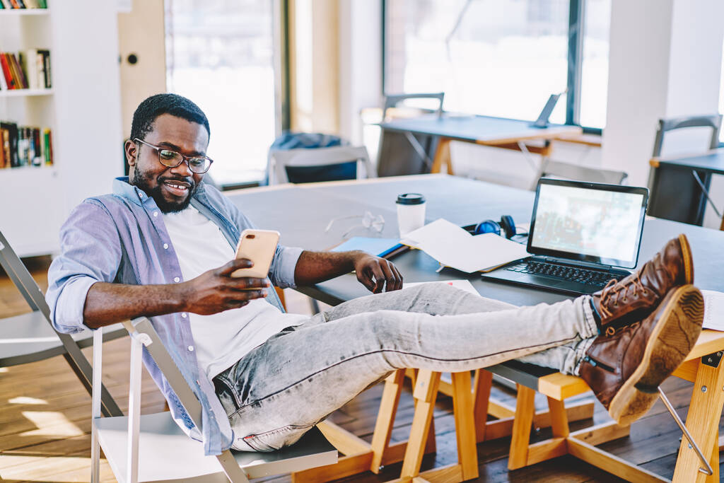 Веселий оптимістичний афроамериканець у окулярах сидить у кріслі з ногами на столі і переглядає мобільний телефон за столом з ноутбуком. - Фото, зображення