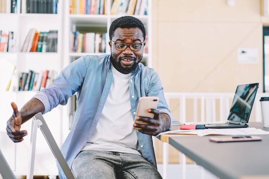 Афроамериканский смарт-мужчина в очках и повседневной одежде смотрит на мобильный телефон, сидя в кресле за столом с ноутбуком - Фото, изображение