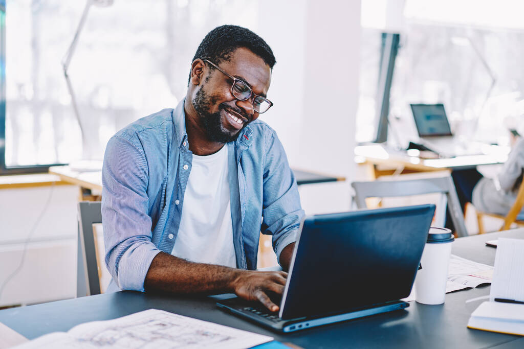 Афроамериканець у окулярах працює за ноутбуком і з радістю посміхається, сидячи за столом біля великих вікон і стопів. - Фото, зображення