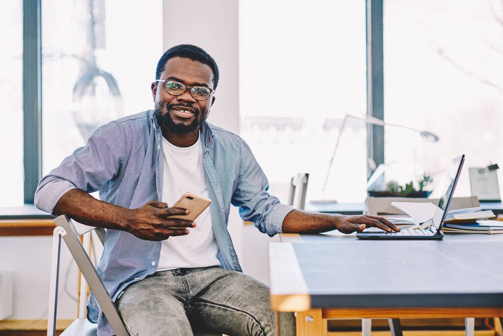 Счастливый афроамериканец со смартфоном в руках сидит за современным рабочим столом с ноутбуком и улыбается в камеру  - Фото, изображение