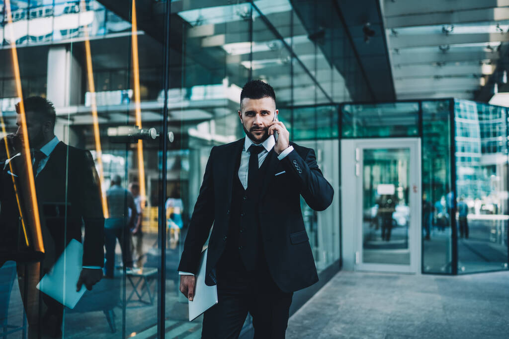 Fókuszált szakállas fickó öltönyben nézi a kamerát, és okostelefonnal beszél, miközben a kortárs irodaház előtt áll, üvegfallal - Fotó, kép