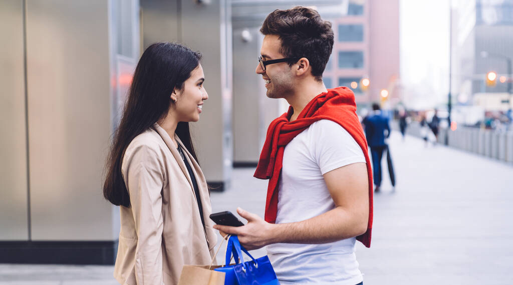 スマートフォンをサーフィンしながら、買い物袋で都市の建物の近くに立っている間、カジュアルな服装で甘い愛情の多様なカップルの側面図や背景にお互いを見て - 写真・画像