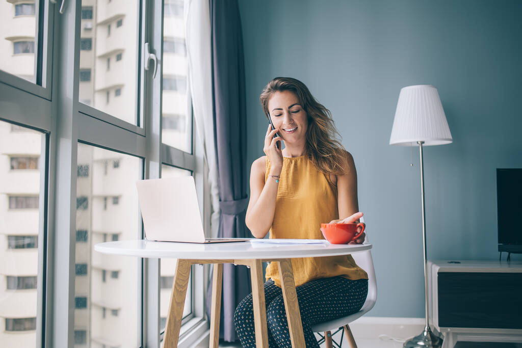 Šťastná mladá žena ve žluté halence bez rukávů sedí u bílého kulatého stolu s notebookem a červeným hrnkem komunikující na smartphone doma - Fotografie, Obrázek