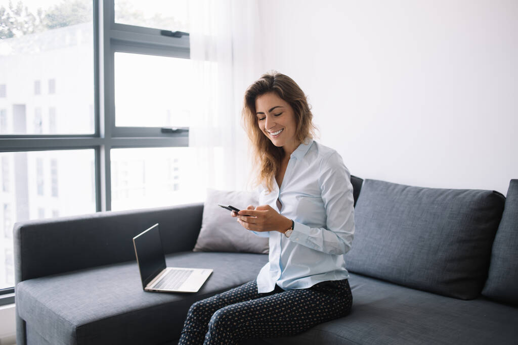 シャツを着た若いビジネスの女性とズボンの上で電話でチャットソファに座ってアパートのリビングルームの近くのコンピュータと笑顔 - 写真・画像
