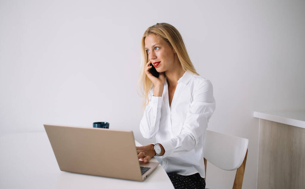 Mujer rubia joven concentrada sentada en la mesa blanca con taza azul en blusa hablando en smartphone usando el portátil en casa y mirando hacia otro lado - Foto, imagen