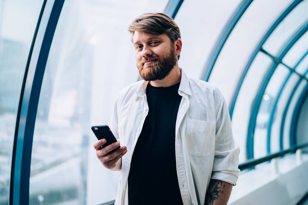 Halve lengte portret van knappe hipster blogger gekleed in casual wit shirt met mobiele telefoon gadget in de hand en kijkend naar de camera tijdens de vrije tijd, concept van technologie en communicatie - Foto, afbeelding