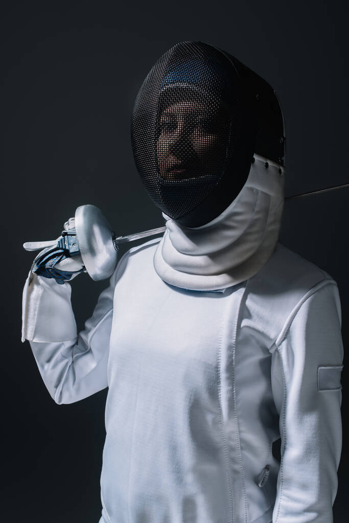 Fencer in fencing mask looking at camera and holding rapier isolated on black  - Photo, Image
