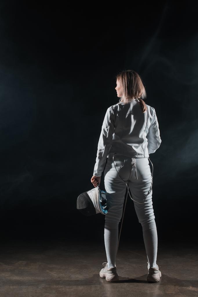 Back view of swordswoman holding fencing mask and rapier on black background with smoke  - Photo, Image