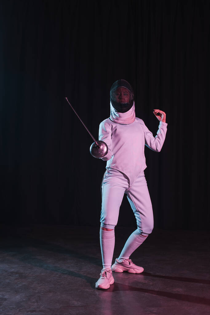 Fencer in fencing mask and suit holding rapier on black background - Photo, Image