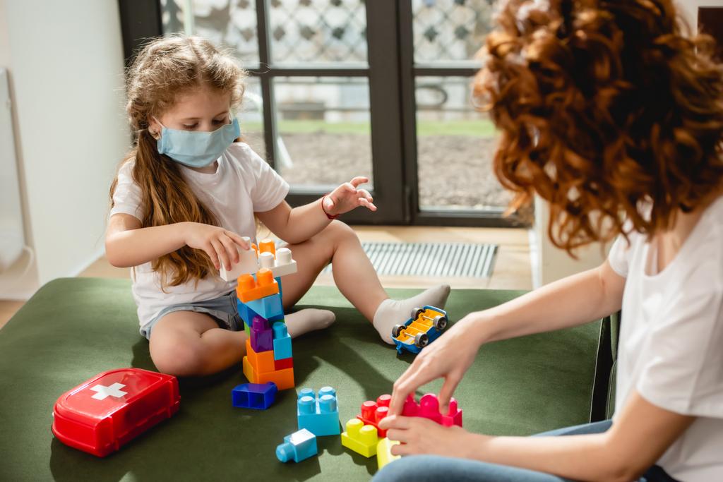 selective focus of cute kid in medical mask playing building blocks with redhead mother  - Photo, Image