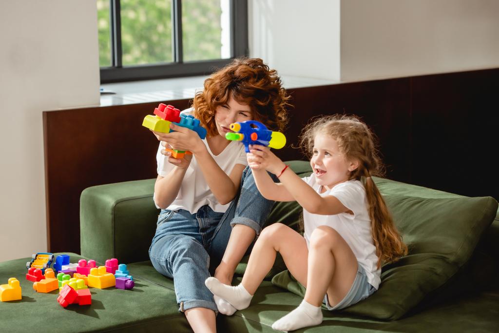 lockige Mutter und Tochter mit Wasserpistole spielen im Wohnzimmer  - Foto, Bild