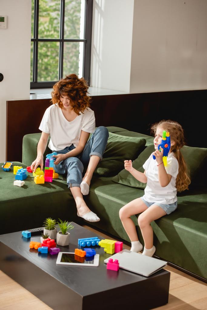 linda hija sosteniendo pistola de agua cerca de la madre y gadgets en sala de estar
  - Foto, imagen