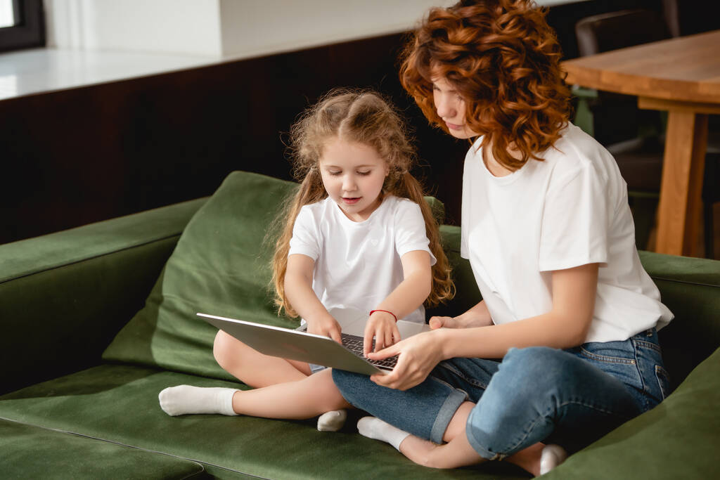 pelirroja madre sosteniendo portátil cerca adorable hija en sala de estar
  - Foto, Imagen