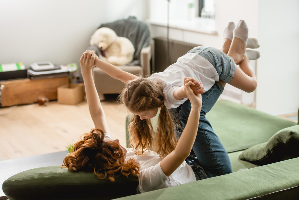 curly mother lying on sofa, lifting cute kid and holding hands in living room  - Photo, Image