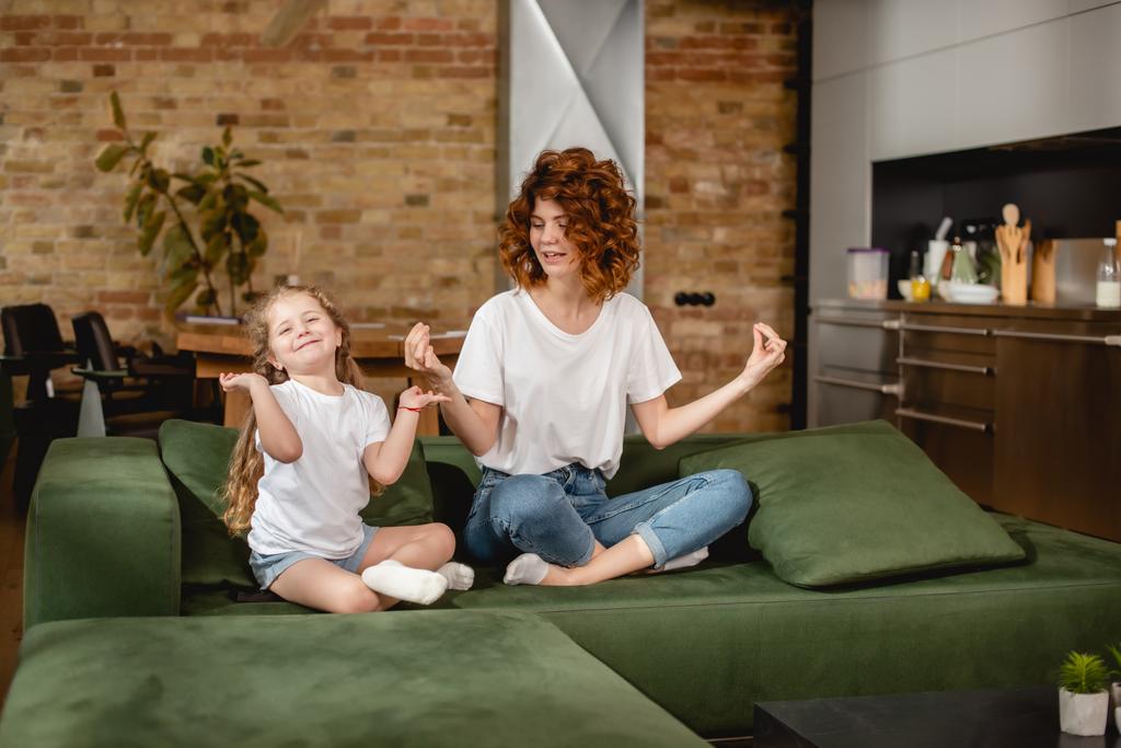 curly mother looking at daughter sitting in lotus pose on sofa - Photo, Image