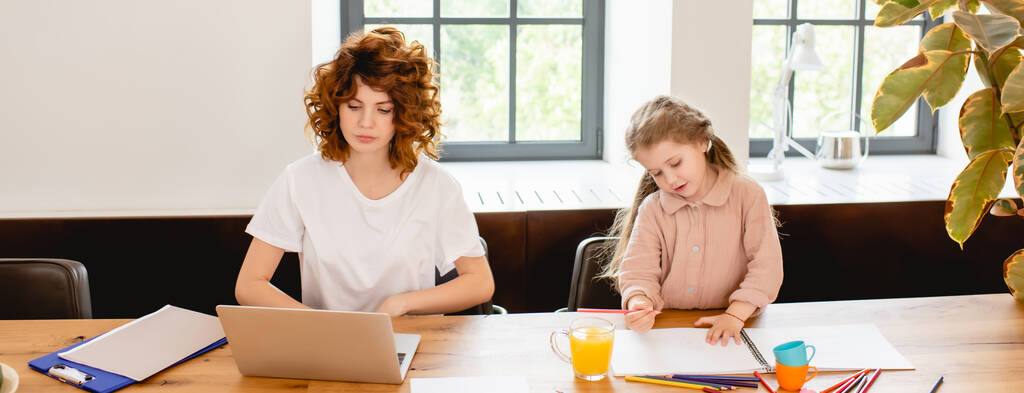 colheita panorâmica de mãe freelancer encaracolado usando laptop perto de criança desenho em casa
 - Foto, Imagem
