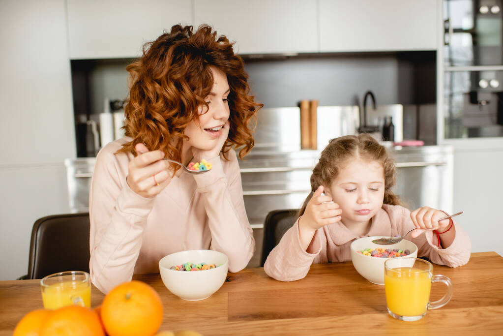 madre rizada y adorable niño sosteniendo cucharas cerca de cuencos con copos de maíz y vasos de jugo de naranja
  - Foto, imagen
