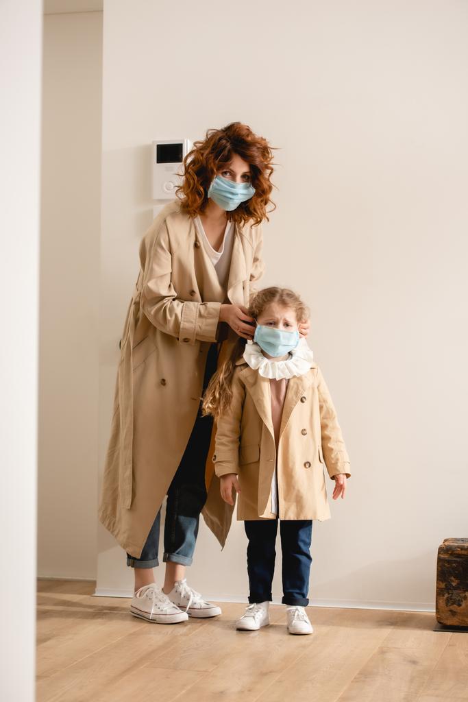lockige Mutter im Trenchcoat mit medizinischer Maske auf süßer Tochter  - Foto, Bild