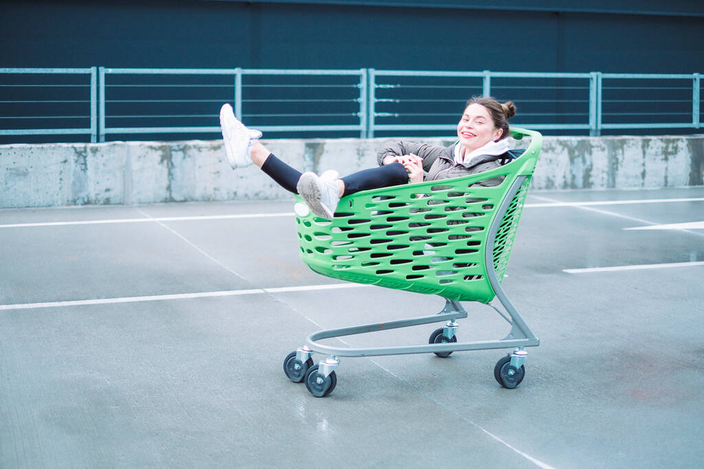 Une dame aux cheveux bruns mince, vêtue d'une tenue décontractée, s'amuse avec un chariot d'épicerie près du centre commercial
. - Photo, image