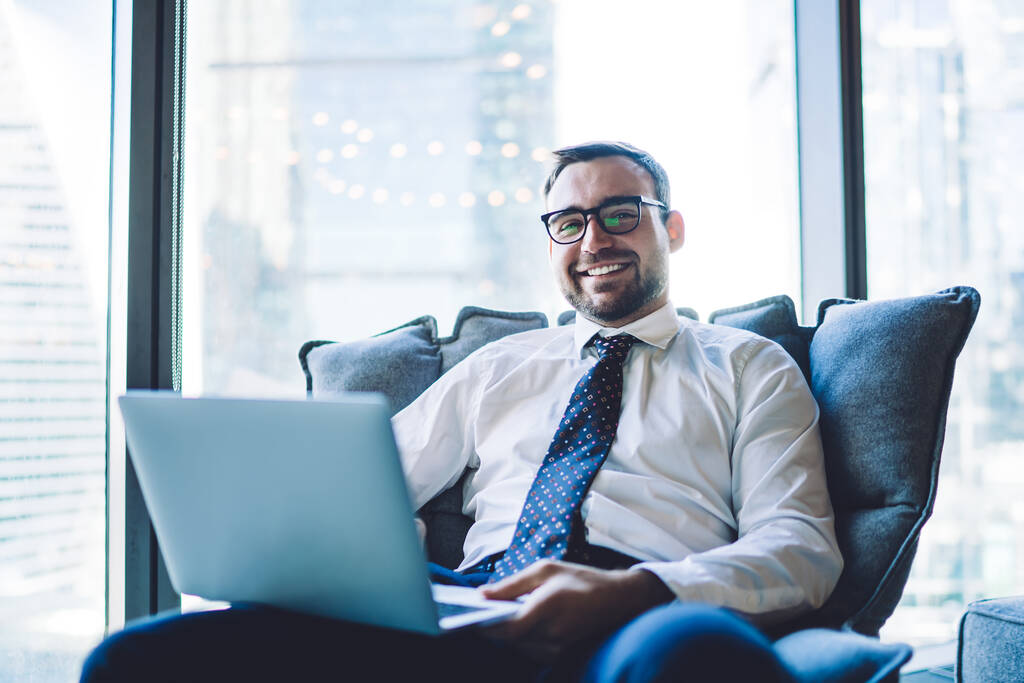 Cheerful macho sonriente con traje formal con corbata usando laptop que comprueba el progreso del proyecto sentado en sillón en la moderna sala de oficina. - Foto, imagen