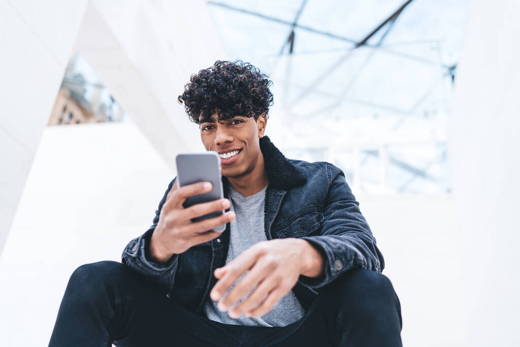 Von unten glücklicher junger afroamerikanischer Mann in stilvoller Kleidung mit lockigem Haar, der sein Smartphone benutzt und in die Kamera schaut, während er in einem weißen modernen Gebäude sitzt - Foto, Bild
