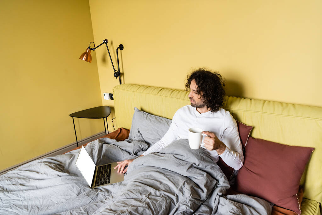 vista de ángulo alto del joven freelancer utilizando el ordenador portátil con pantalla blanca mientras sostiene la taza de café en la cama
 - Foto, Imagen