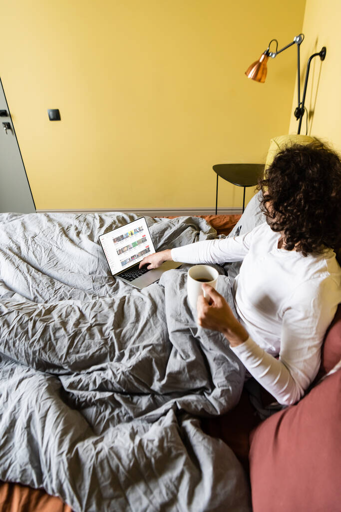 KYIV, UKRAINE - APRIL 25, 2020: high angle view of curly freelancer using laptop with Youtube website while holding cup of coffee in bed - Photo, Image
