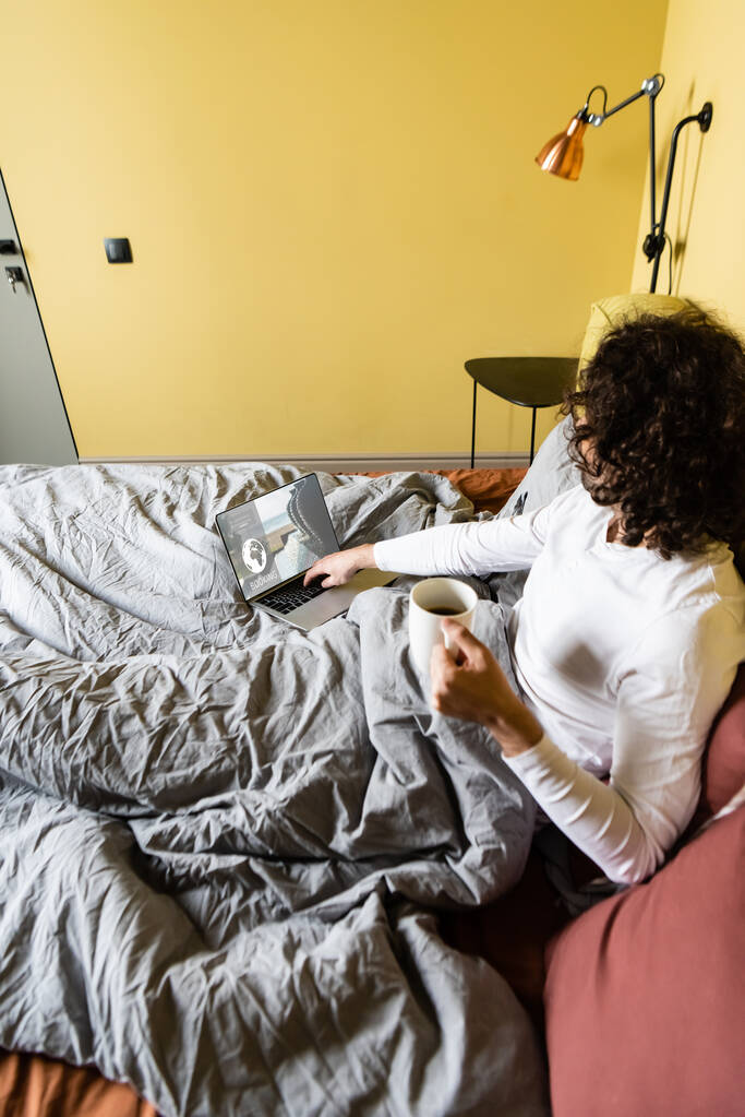 vista de ángulo alto del freelancer rizado utilizando el ordenador portátil con entradas sitio web en línea mientras sostiene la taza de café en la cama
 - Foto, Imagen