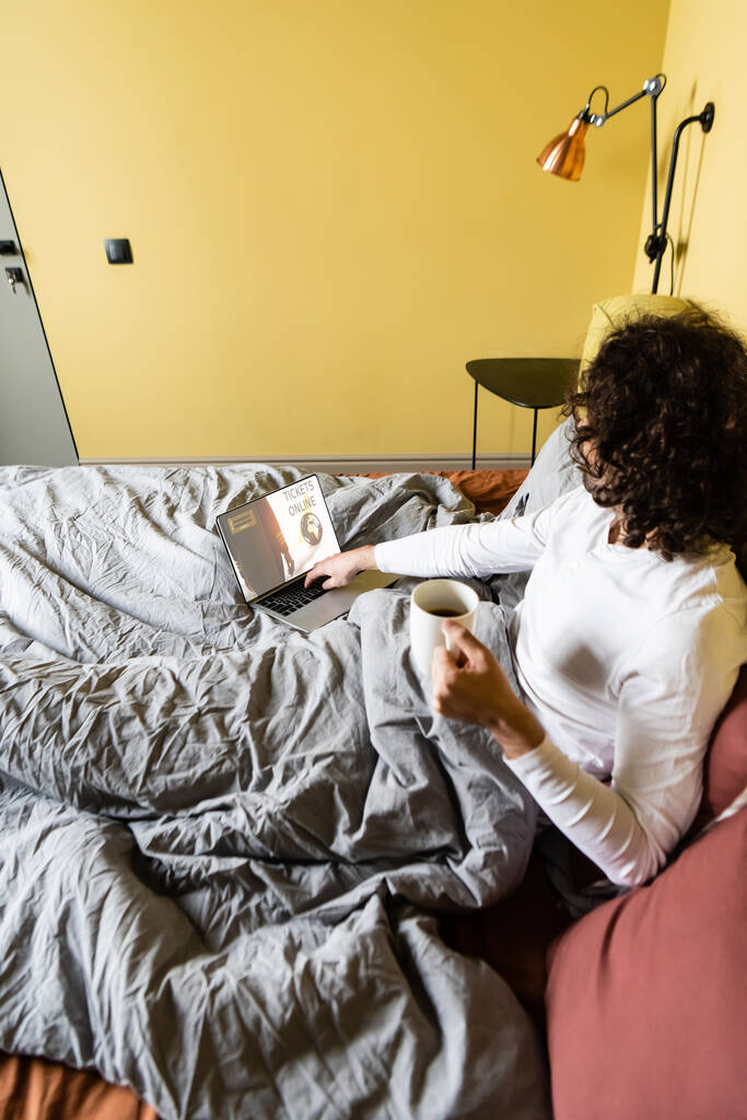 high angle view of curly freelancer holding cup of coffee and using laptop with tickets online website in bed - Photo, Image