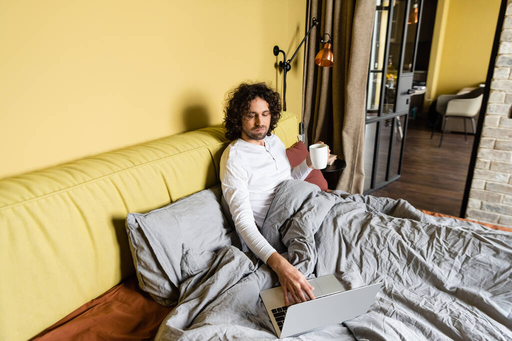Freelancer encaracolado usando laptop enquanto segurando xícara de café na cama
 - Foto, Imagem
