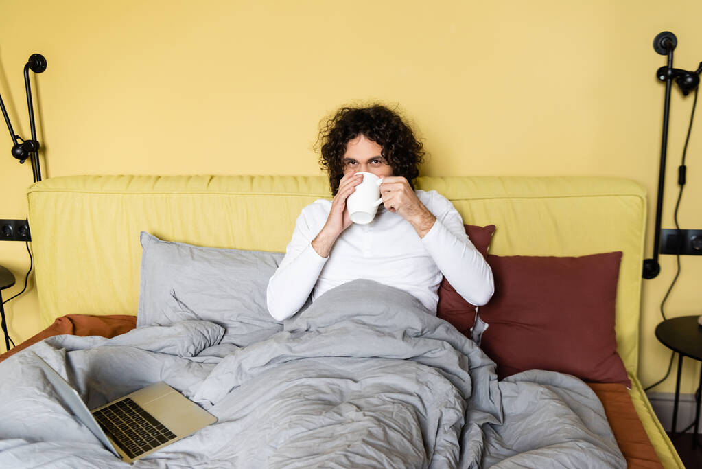 krullend freelancer drinken van koffie in bed in de buurt van laptop en kijken naar camera - Foto, afbeelding