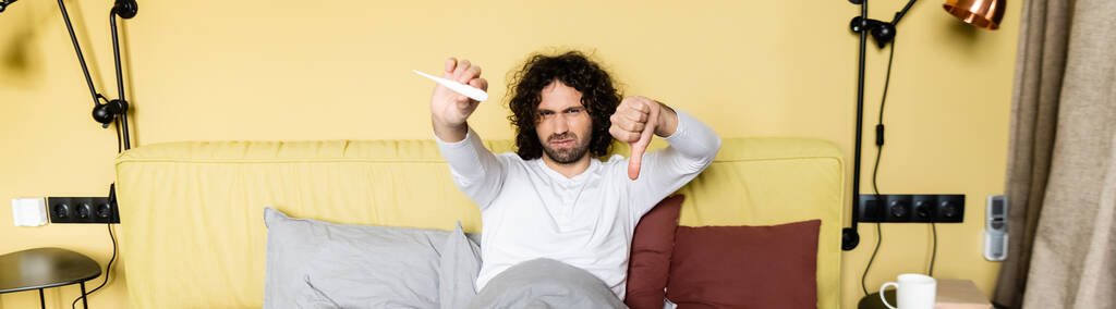 vízszintes kép elégedetlen férfi tartja hüvelykujját lefelé, és nézi a hőmérő az ágyban - Fotó, kép