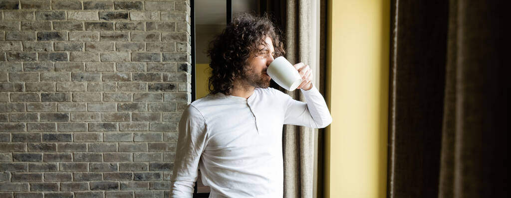 горизонтальне зображення красивого чоловіка в піжамі п'є каву, дивлячись у вікно вранці
 - Фото, зображення