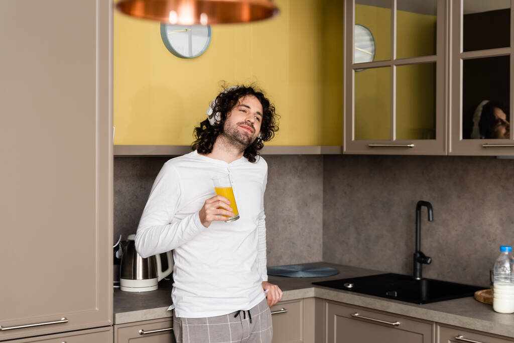 χαμογελαστός άνθρωπος με ασύρματα ακουστικά κοιτάζοντας μακριά, ενώ κρατώντας χυμό πορτοκάλι - Φωτογραφία, εικόνα