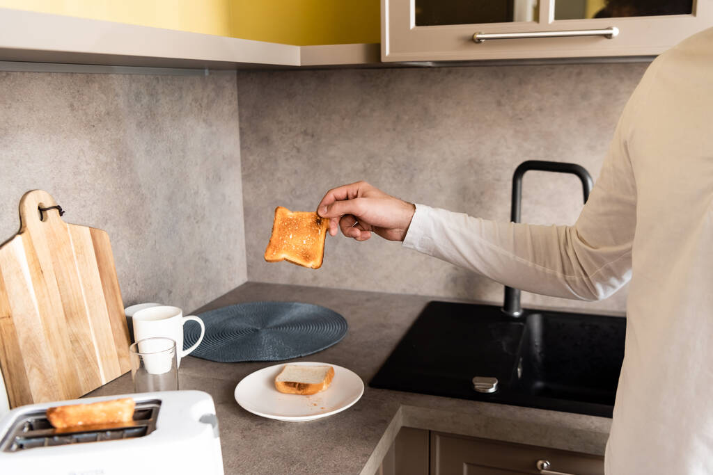 abgeschnittene Ansicht eines Mannes, der leckeres frittiertes Brot in der Nähe eines Toasters in der Küche hält - Foto, Bild