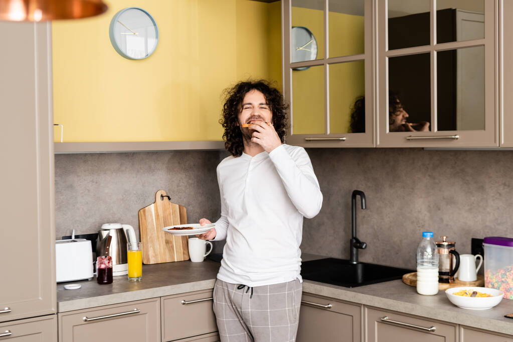 χαρούμενη, σγουρά άνθρωπος τρώει νόστιμα τοστ με μαρμελάδα, ενώ κοιτάζοντας κάμερα - Φωτογραφία, εικόνα