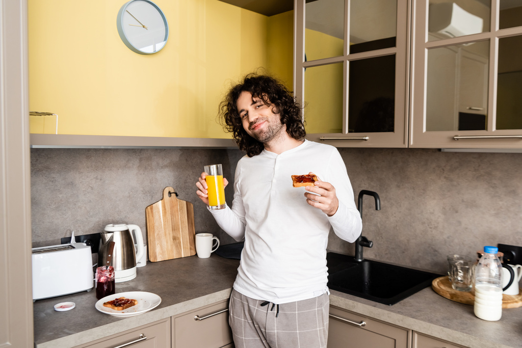 カメラで微笑みながらオレンジジュースとトーストを持ったパジャマ姿の幸せな男 - 写真・画像