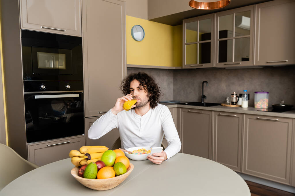 handsome man looking at camera while drinking orange juice and using smartphone during breakfast - Photo, Image