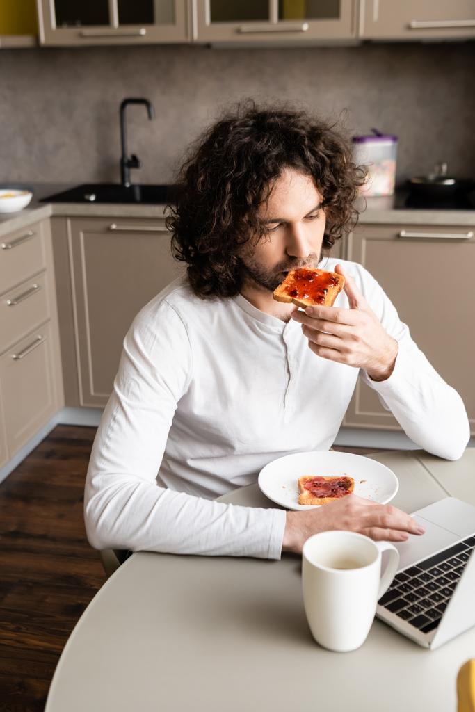 στοχαστικός ελεύθερος επαγγελματίας τρώει τοστ, ενώ κοιτάζοντας φορητό υπολογιστή κοντά σε φλιτζάνι καφέ - Φωτογραφία, εικόνα